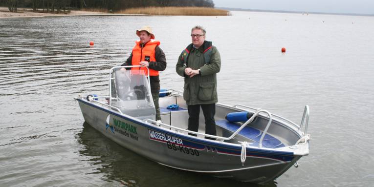 In einem Boot mit einem Rumpf aus korrosionsbeständigem Aluminium stehen zwei Männer und blicken in Richtung Ufer.